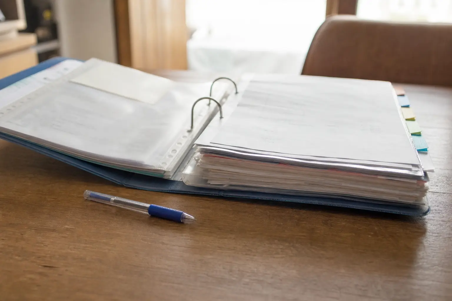 Un classeur ouvert contenant des courriers et des factures séparés par des intercalaires, posé sur une table à côté d'un stylo, dans la lumière du jour