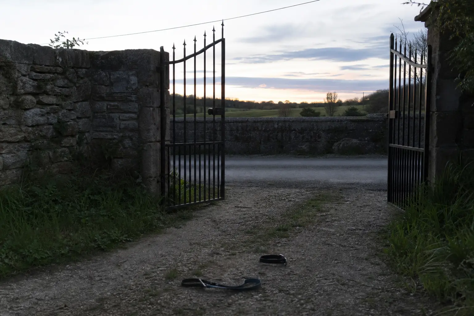 Un portail de jardin entrouvert sur une route de campagne au crépuscule, un collier et une laisse posés au sol devant