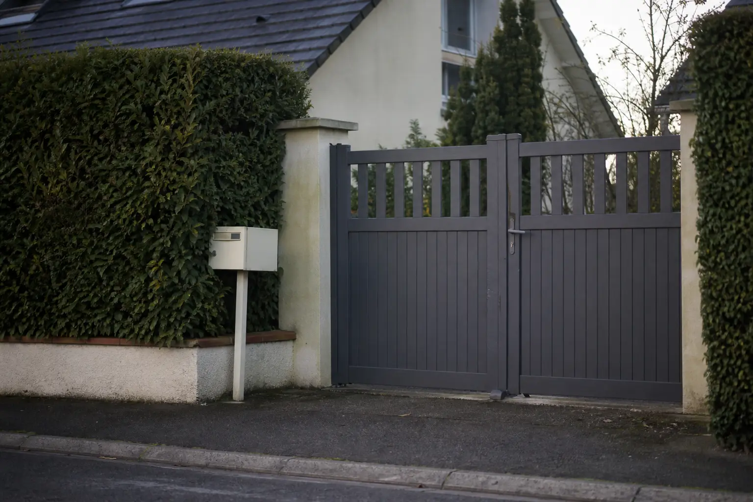 Un portail de jardin fermé vu depuis la rue, avec une haie taillée et une boîte aux lettres placée en limite de propriété