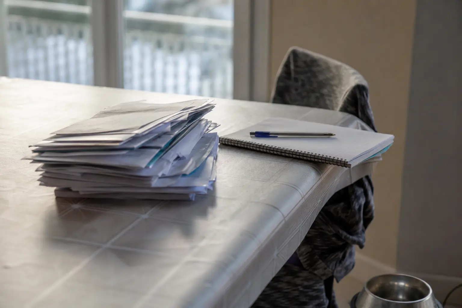 Des factures empilées, un carnet et une gamelle de chien vide posés sur une table de cuisine dans une lumière d'hiver